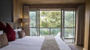 A large bed with a white and beige bedspread, multiple pillows, and a view of trees through large windows. - Menlyn Boutique Hotel (Pretoria)