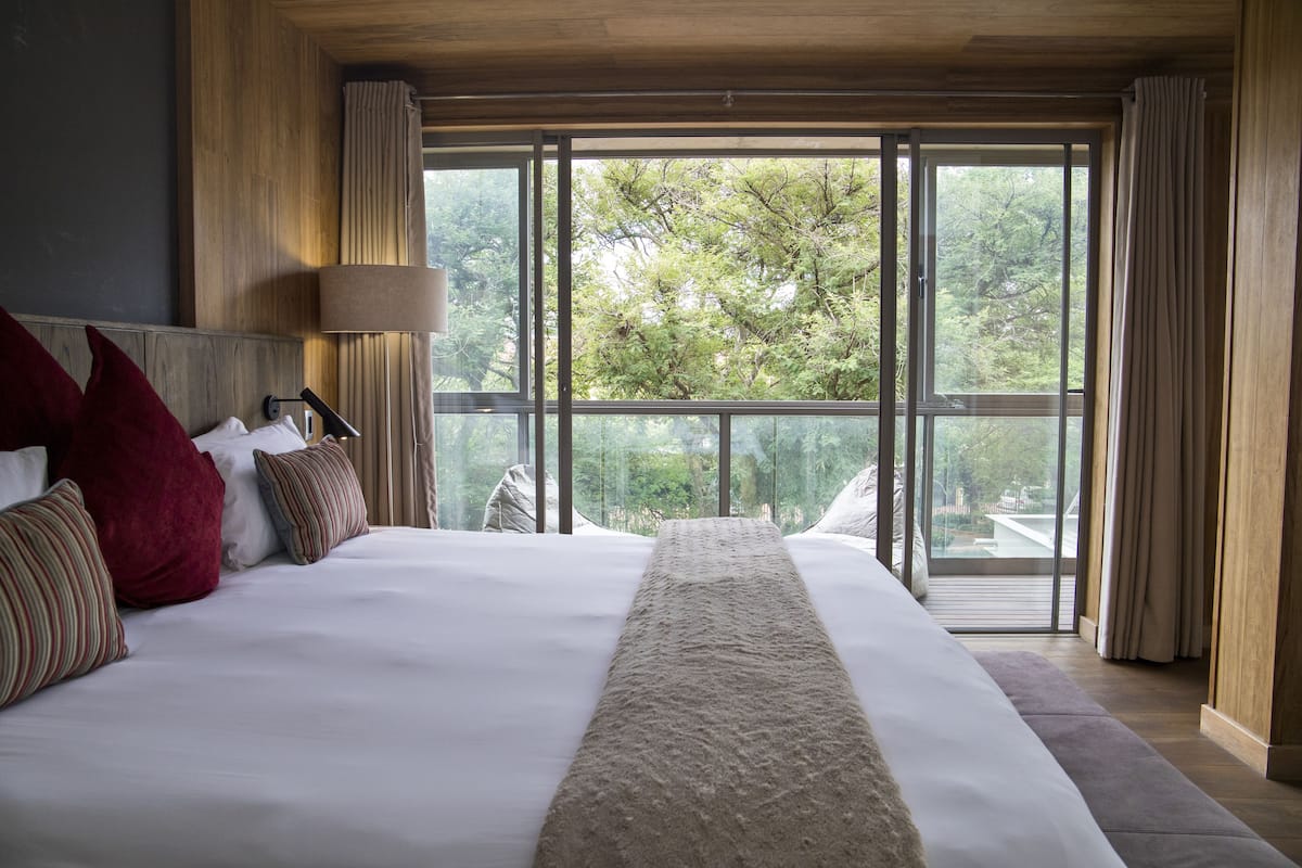 A large bed with a white and beige bedspread, multiple pillows, and a view of trees through large windows.