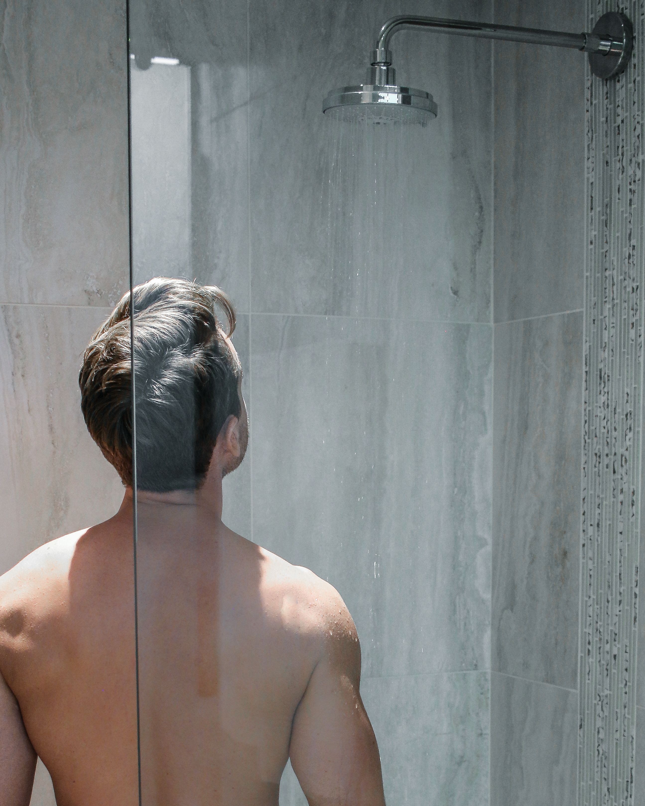 Une personne se trouve dans une douche dotée d’une porte en verre, d’un pommeau de douche et d’un mur carrelé.