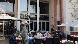 An outdoor dining area with tables and chairs, a statue, and a building with large windows and a covered patio. - Menlyn Boutique Hotel (Pretoria)