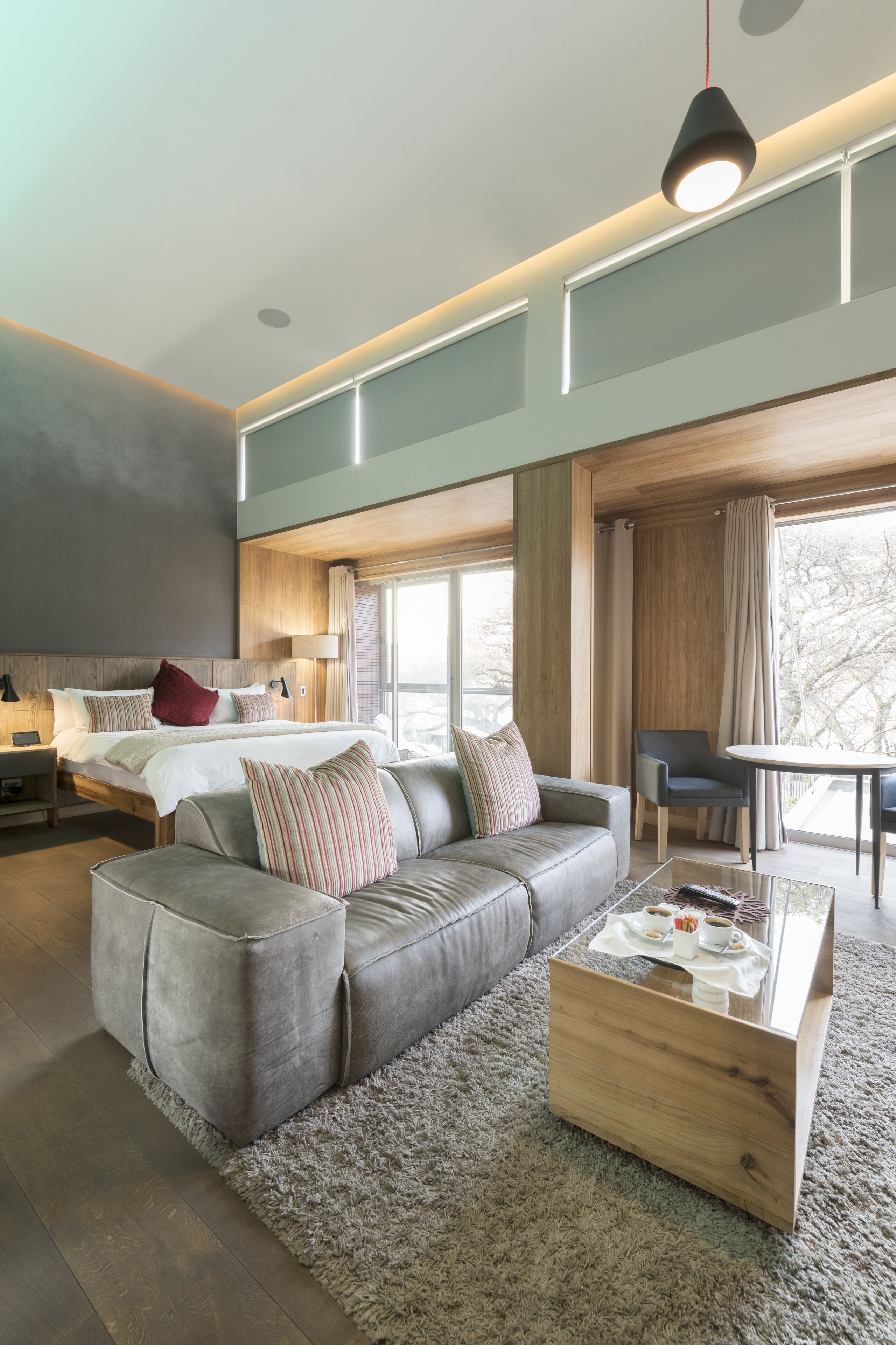 Una habitación de hotel moderna con una cama grande, un sofá gris, una mesa de centro de madera y vistas al exterior.
