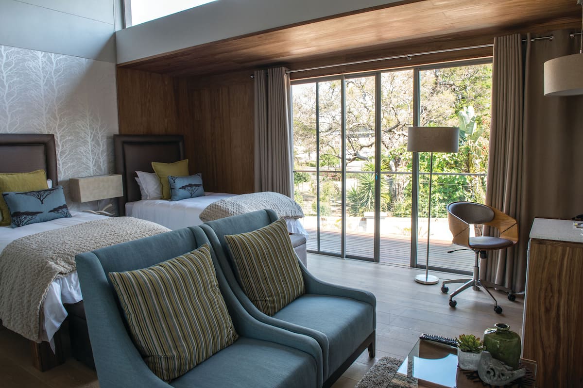 A hotel room with two beds, a sofa, a desk, and a large window with a view of trees.