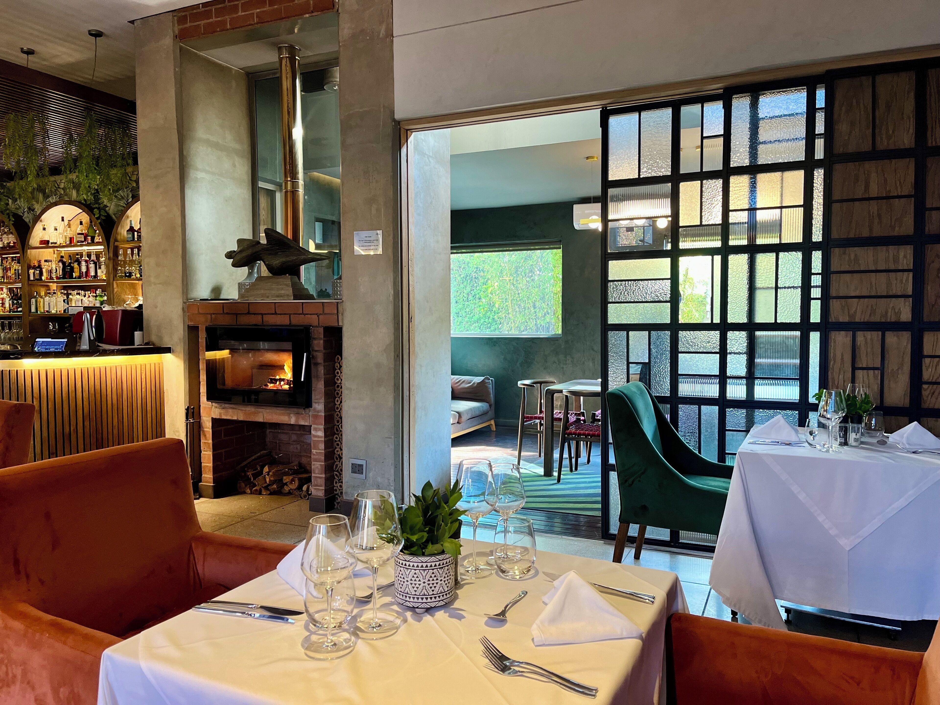 A restaurant with a brick fireplace, a bar area with shelves of bottles, and a view through large windows.