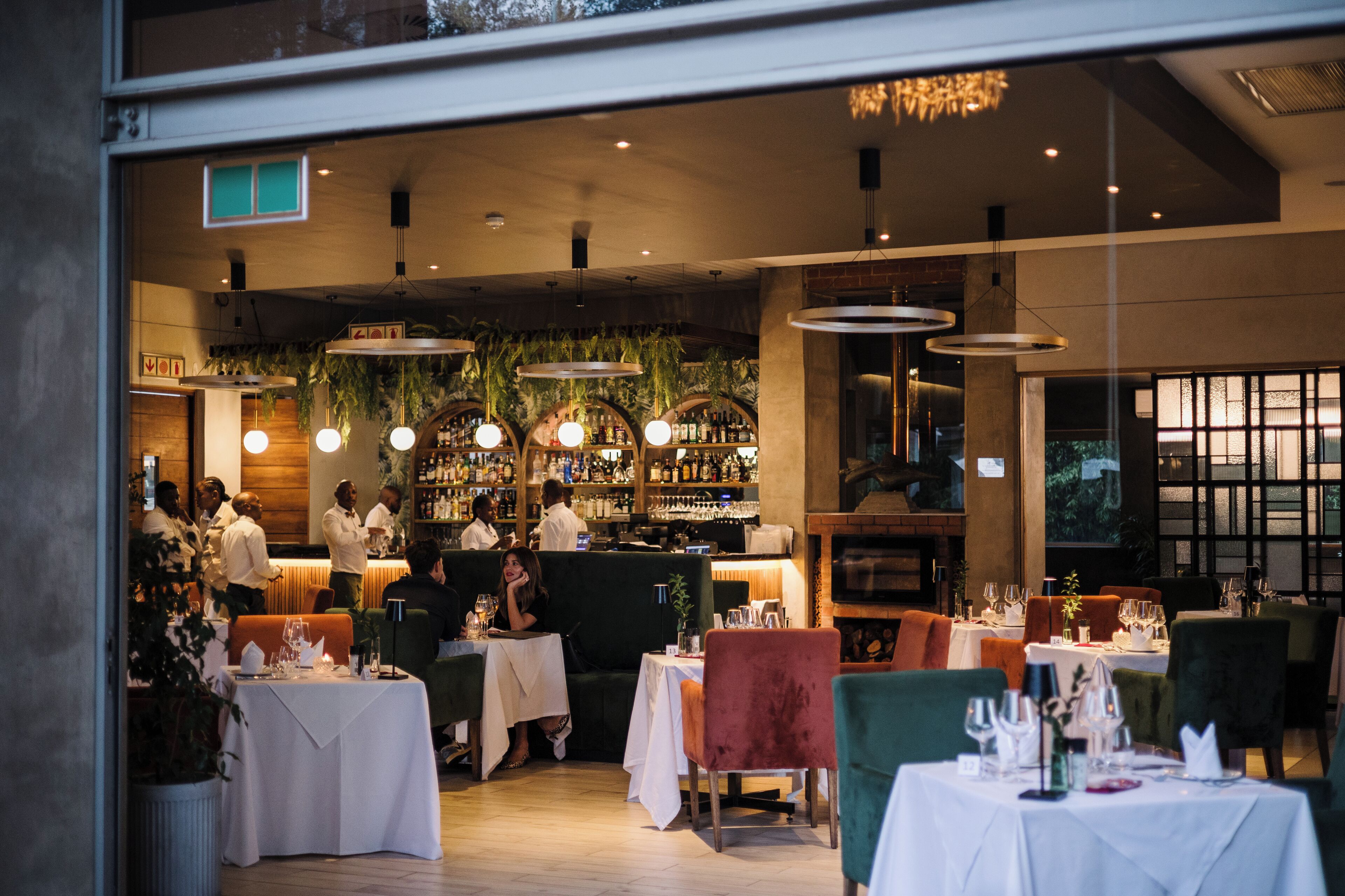 A restaurant with multiple tables and chairs, a bar area with bottles, and hanging plants.