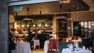A restaurant with multiple tables and chairs, a bar area with bottles, and hanging plants.