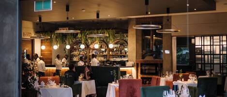 A restaurant with multiple tables and chairs, a bar area with bottles, and hanging plants.