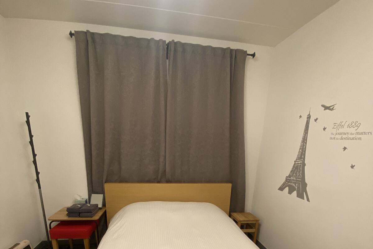 Een slaapkamer met een bed, nachtkastje, lamp, wandklok en een muursticker van de Eiffeltoren.