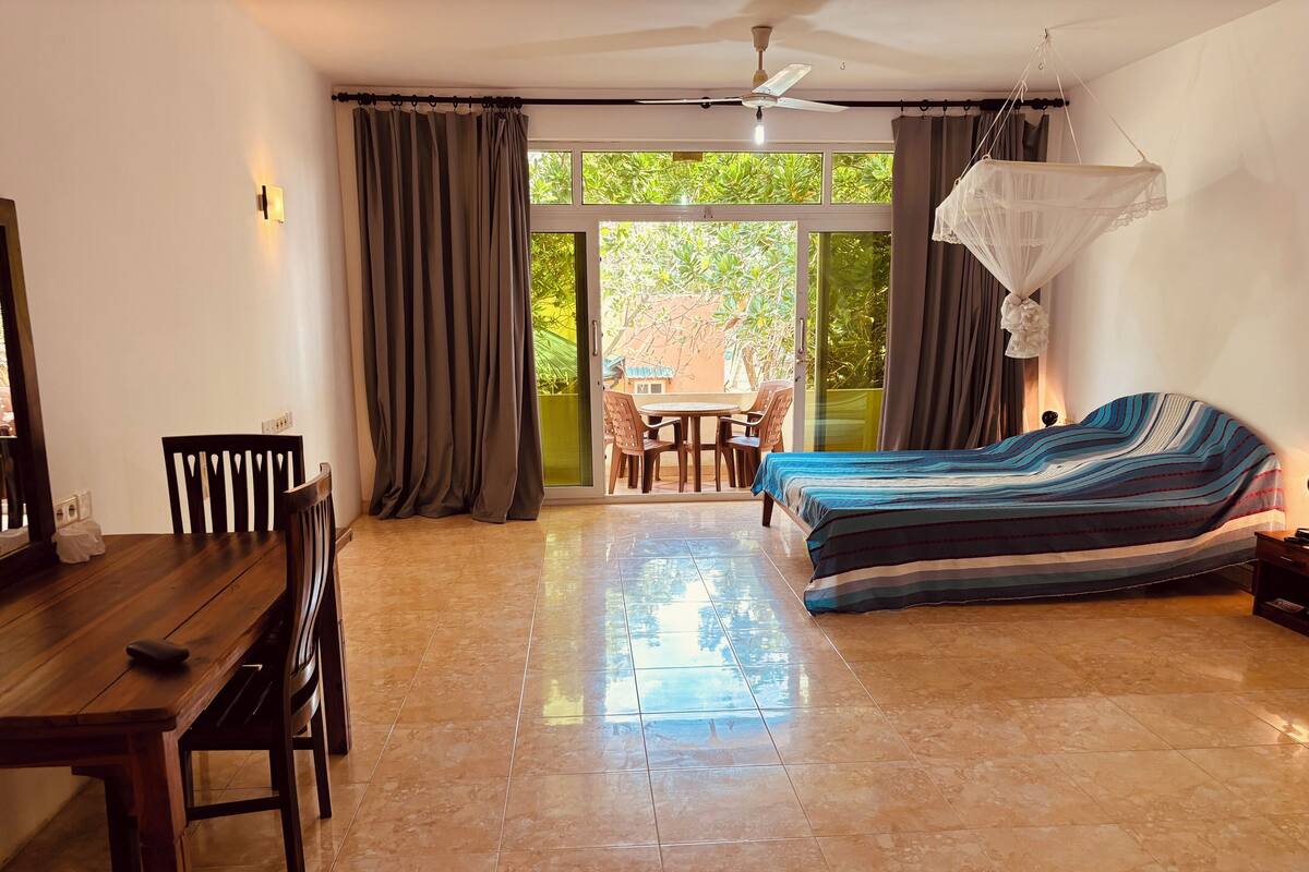 A room with a bed, a desk, and a chair. There is a window with curtains and a view of greenery outside.