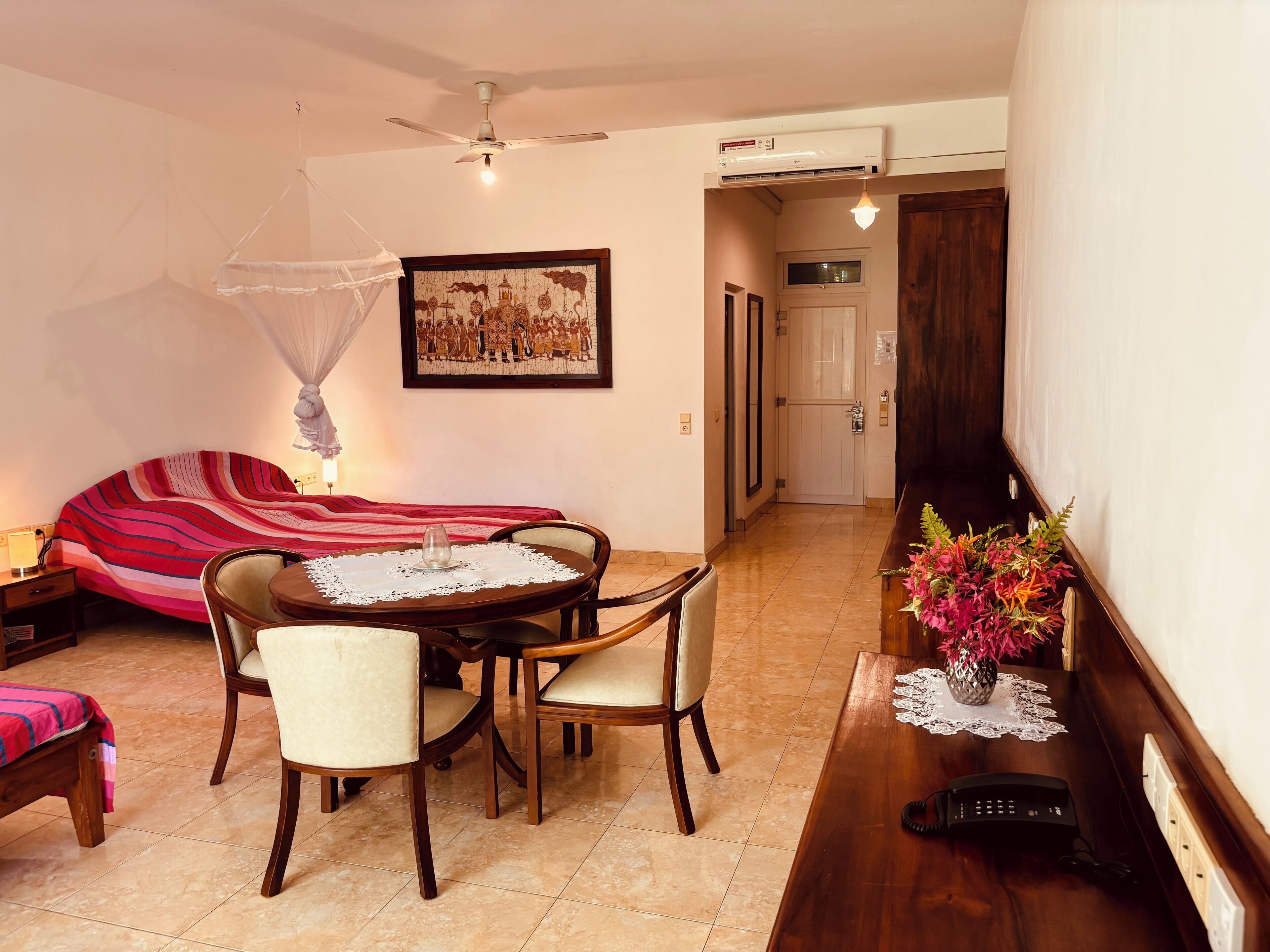 Una habitación con cama, una mesa de comedor con sillas, un ventilador de techo, un equipo de aire acondicionado, una puerta y un aplique en la pared.