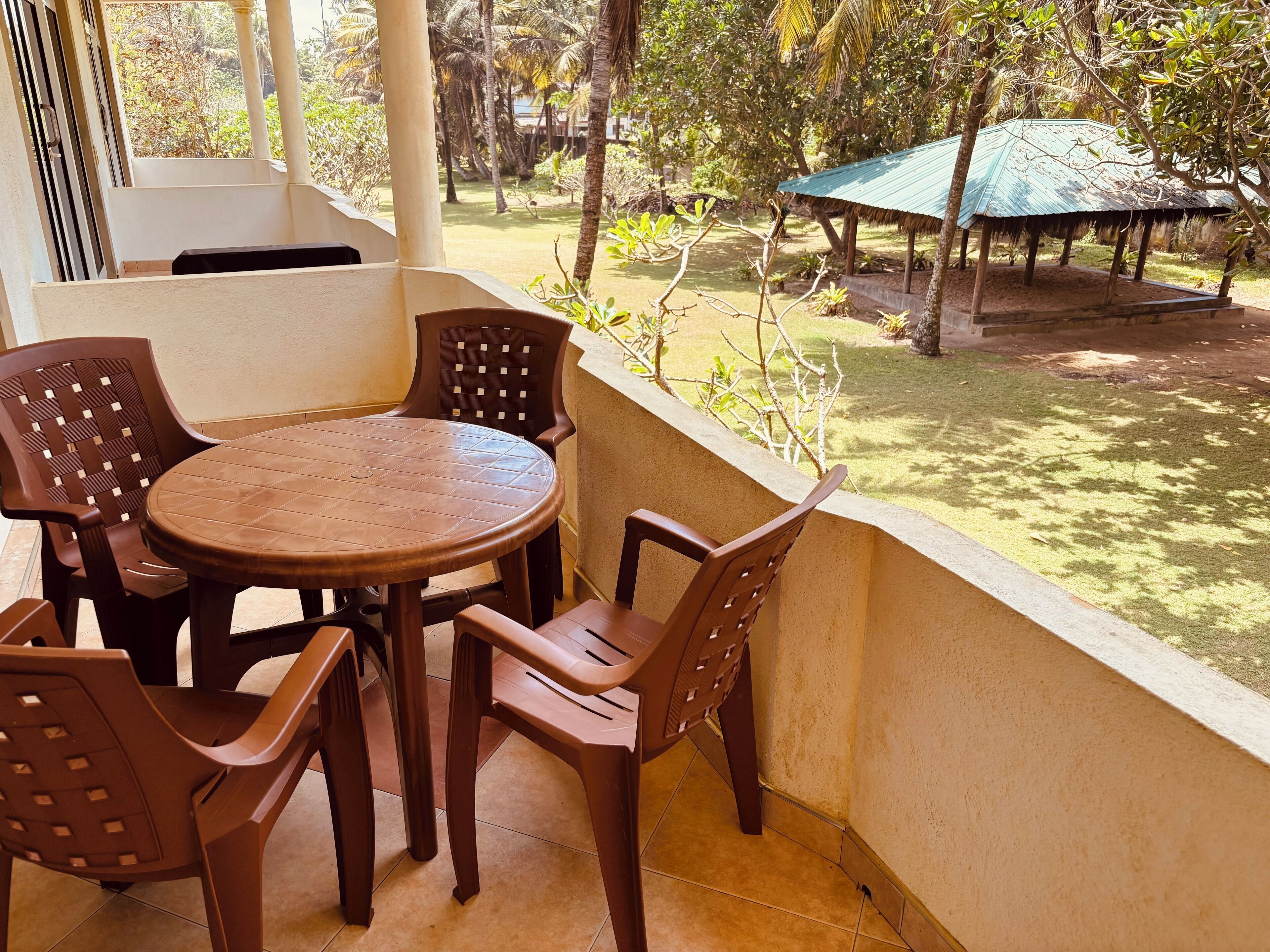 Un balcón con una mesa redonda y cuatro sillas, con vista a un jardín que incluye una estructura con techo de paja.