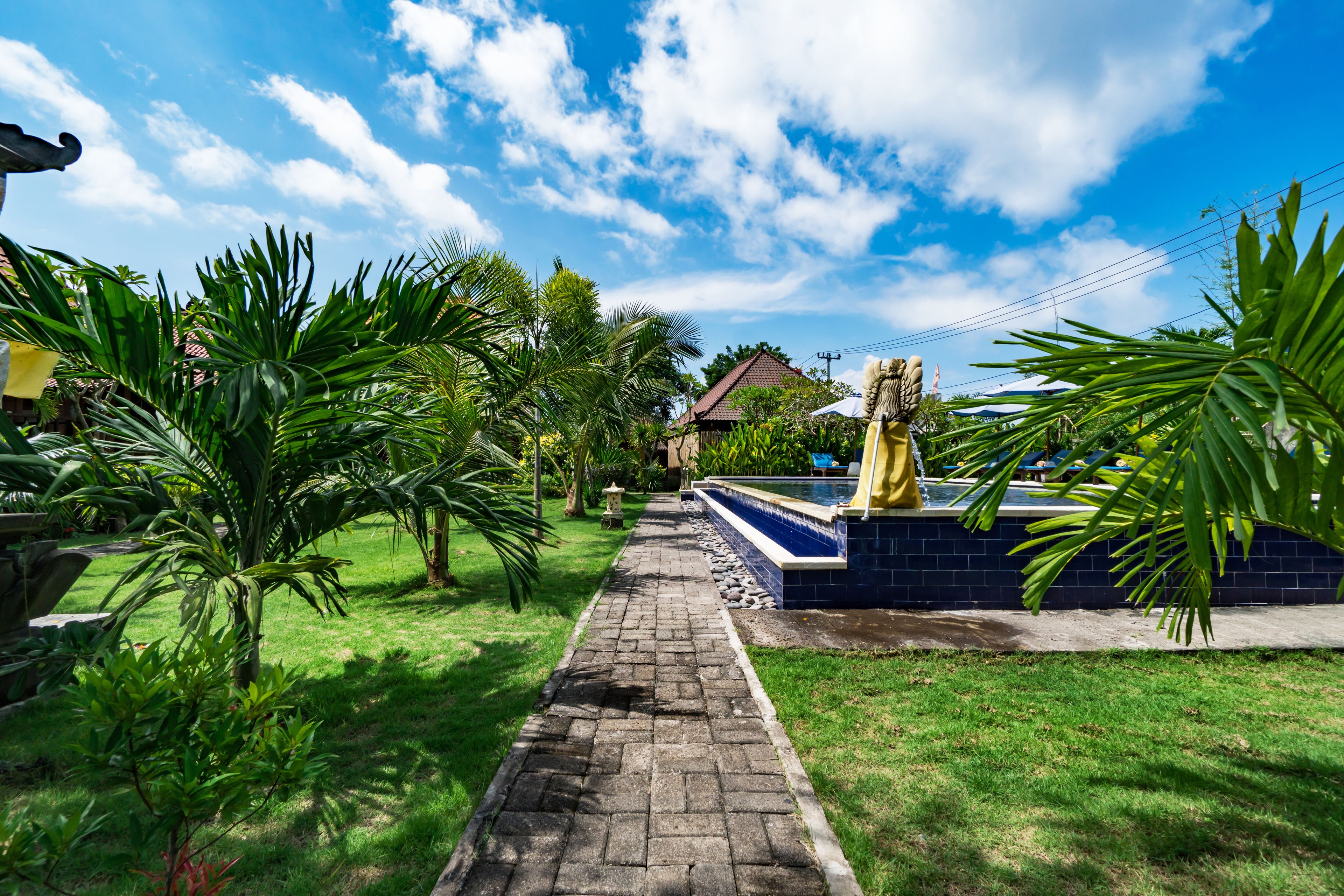 Un sendero de jardín que conduce a una zona de piscina con una estatua, rodeada de abundante vegetación y un cielo azul despejado.
