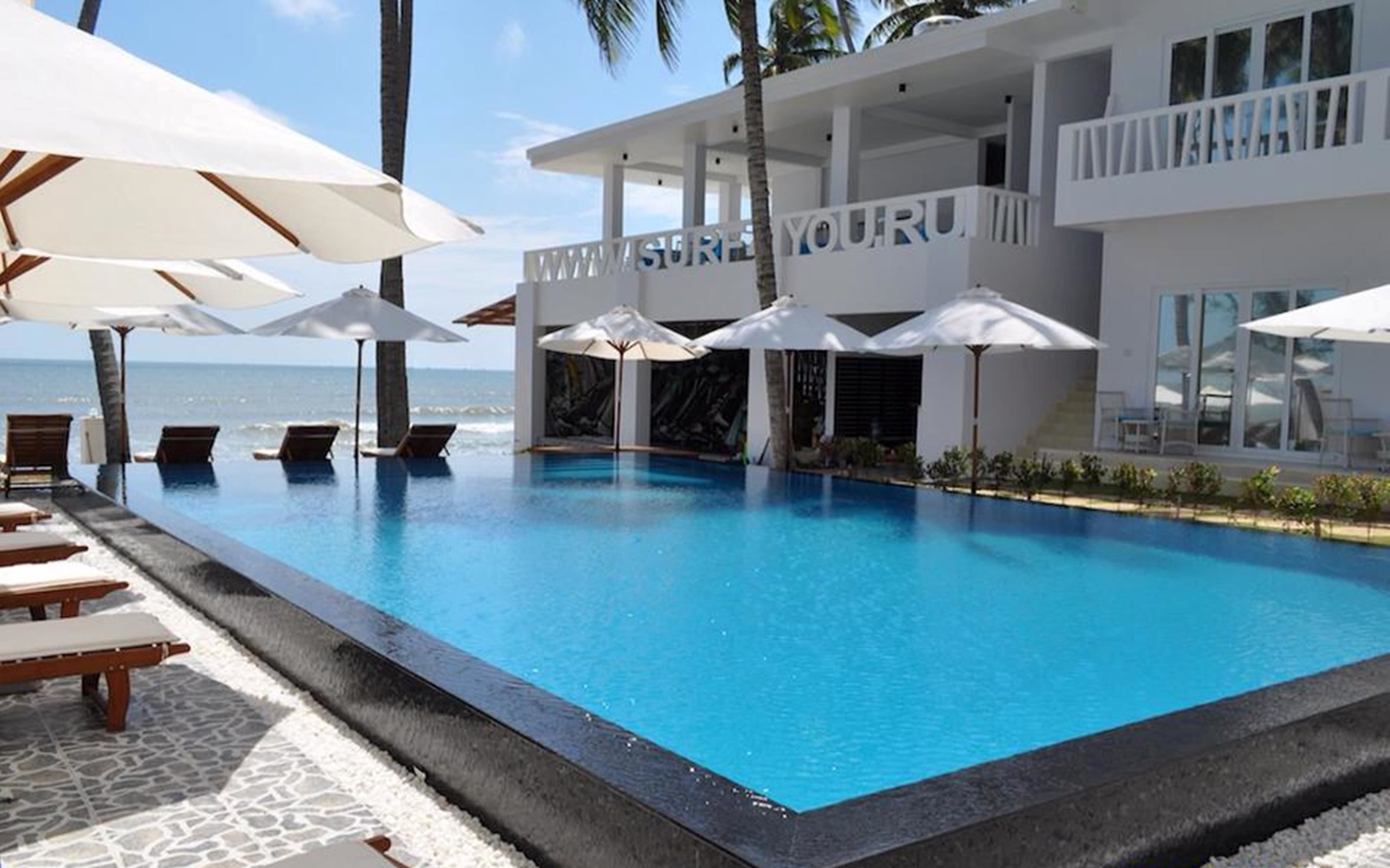 Une piscine avec des chaises longues et des parasols, un bâtiment doté d’un balcon et une vue sur l’océan.