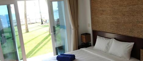 Habitación de hotel con cama, mesita de noche, lámpara y vista a un césped verde y un cuerpo de agua a través de una puerta corrediza de vidrio.
