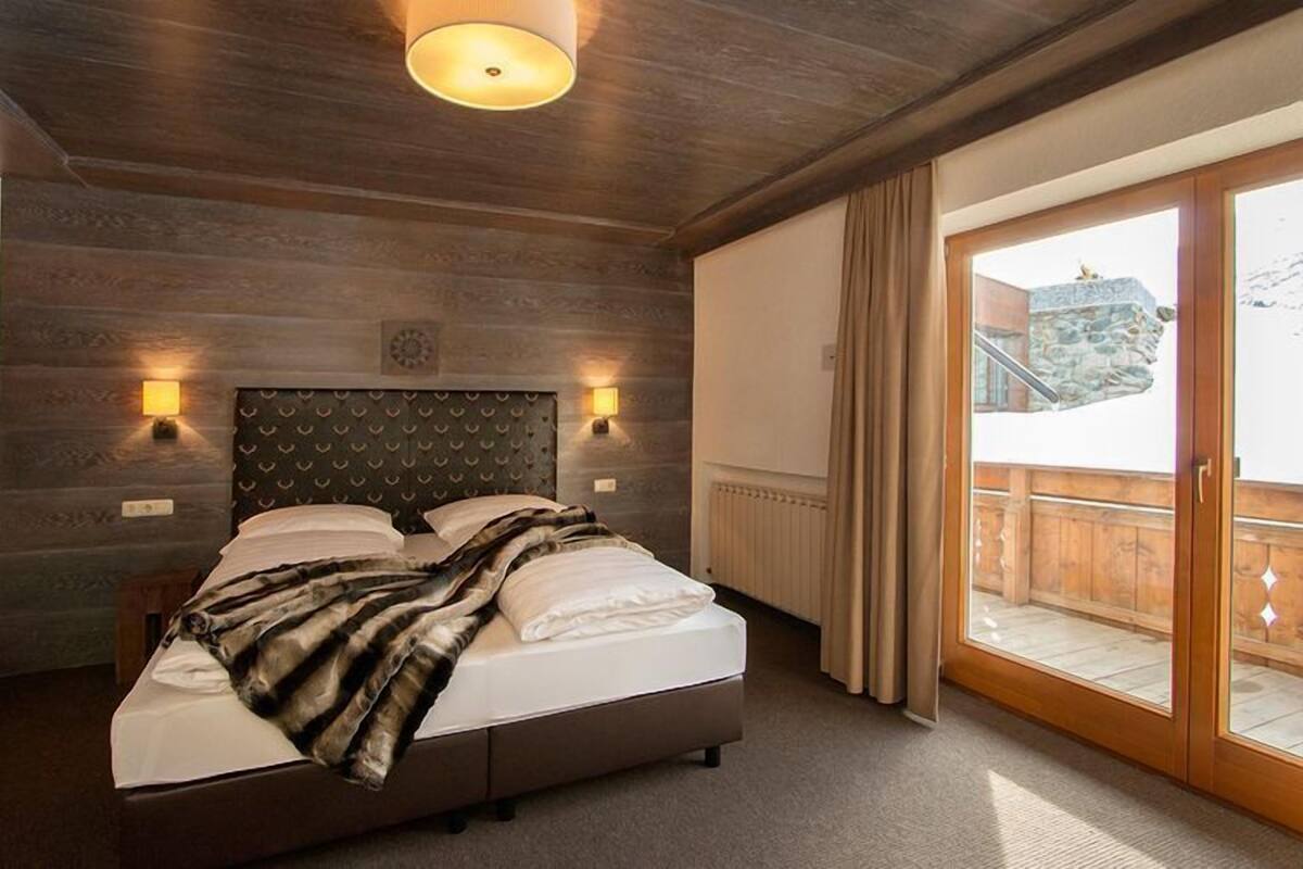 Een slaapkamer met een houten hoofdeinde, een bed met witte lakens, twee wandlampen en uitzicht op een balkon met een stenen muur.