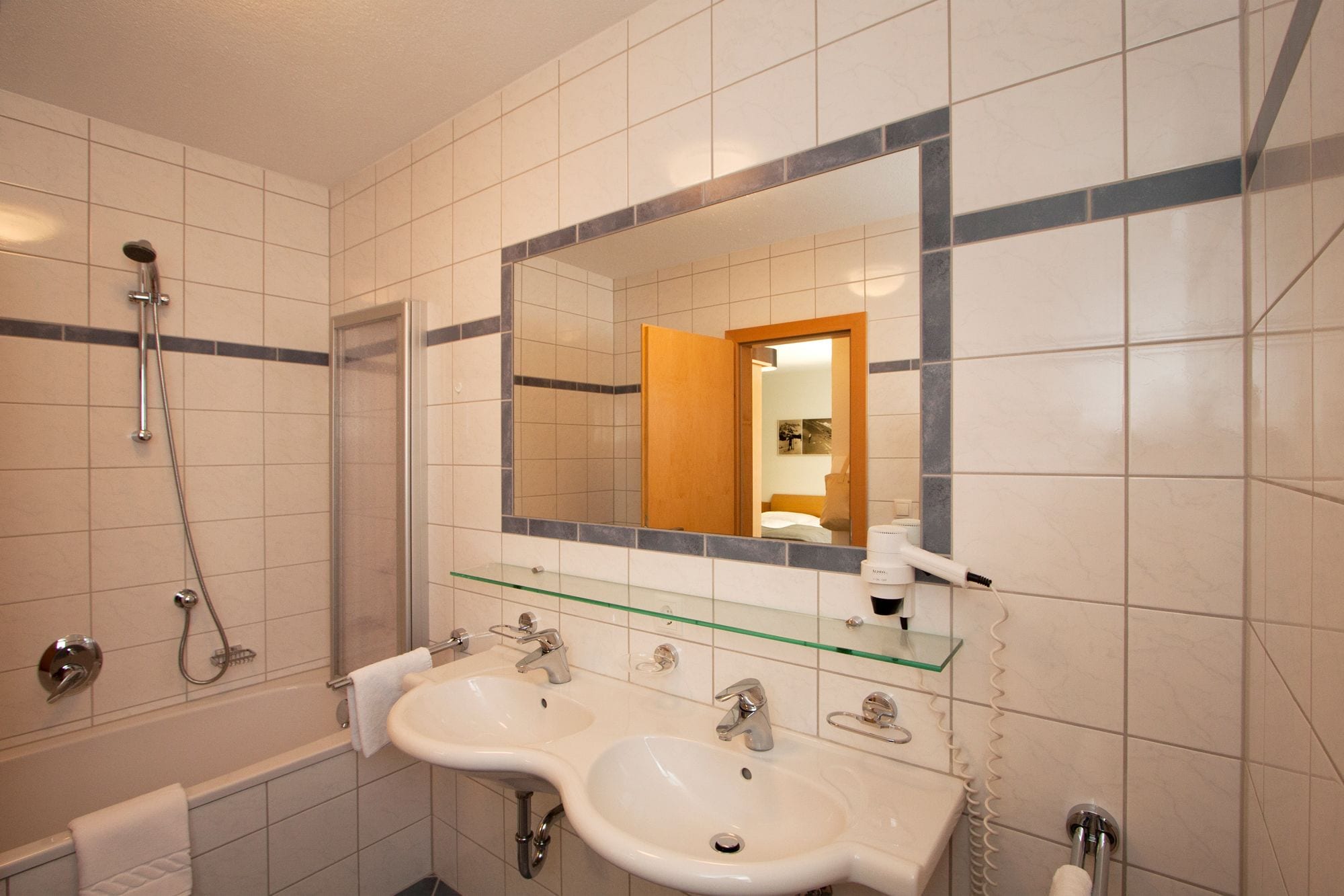 Un baño con dos lavamanos, un estante de vidrio, una ducha y un espejo que refleja una habitación con una cama y una mesita de noche.