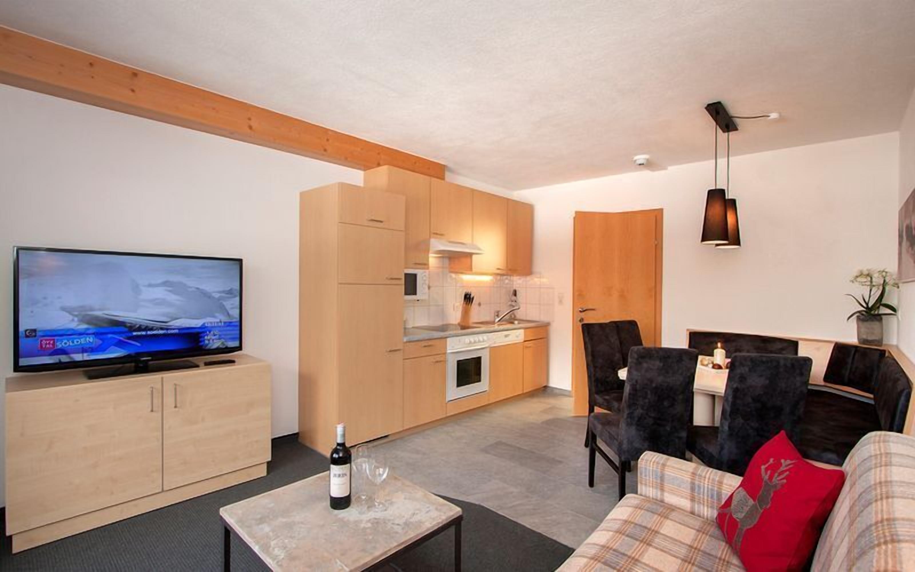 A modern living room with a television, a sofa, a coffee table with a bottle and glasses, and a kitchen area with wooden cabinets and appliances.