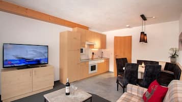 A modern living room with a television, a sofa, a coffee table with a bottle and glasses, and a kitchen area with wooden cabinets and appliances.