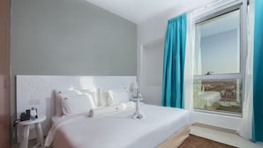 Ein ordentlich bezogenes Bett mit weißen Leinen, ein kleiner Nachttisch mit Toilettenartikeln und ein Fenster mit Vorhängen.
