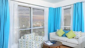 Ein modernes Wohnzimmer mit Blick auf die Stadt, mit einem weißen Sofa, einem gemusterten Sessel und einem kleinen runden Tisch mit Büchern und einer Fernbedienung.