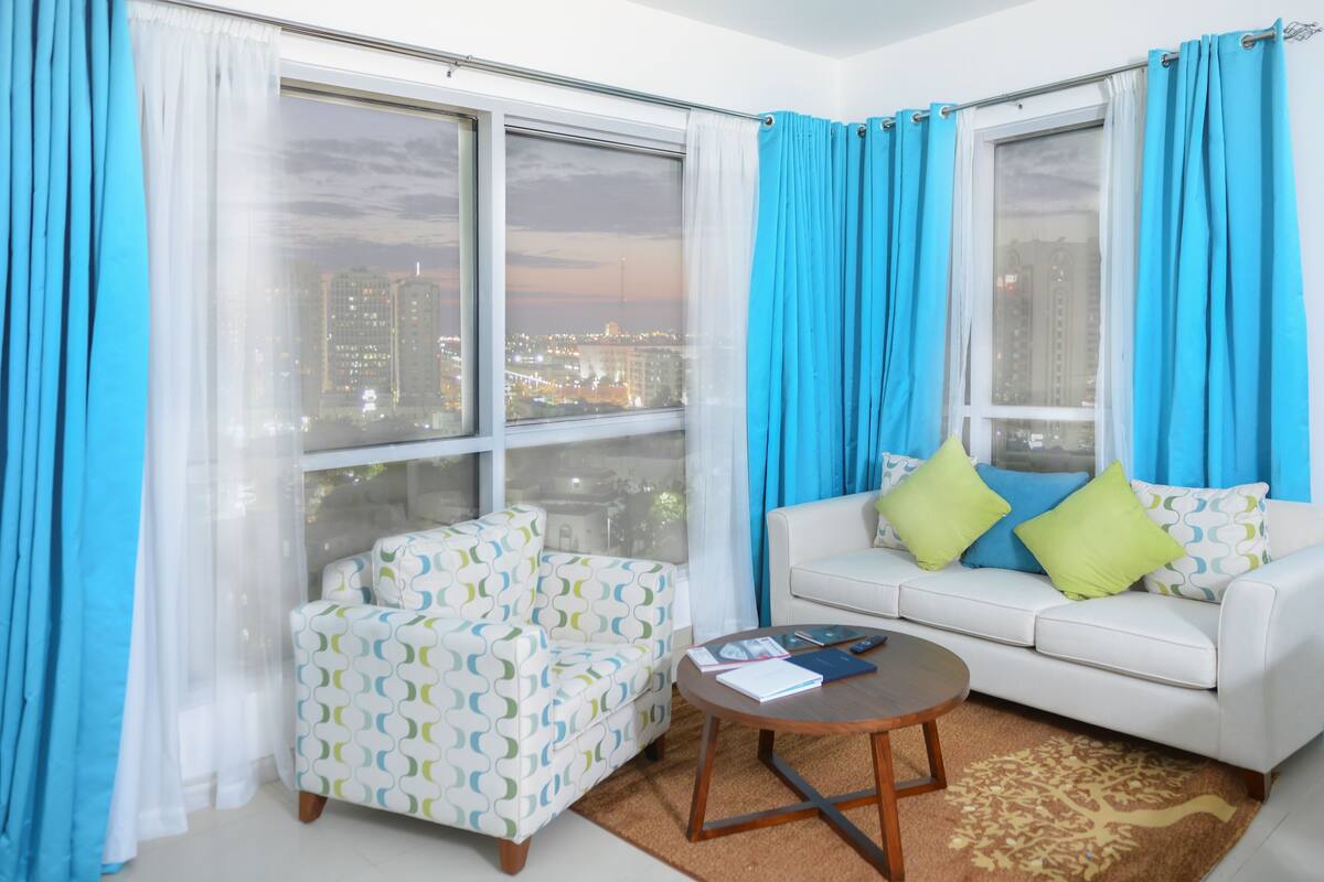 A modern living room with a city view, featuring a white sofa, patterned armchair, and a small round table with books and a remote control.