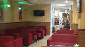 A row of red cushioned seating with a flat-screen TV mounted on the wall, a wooden panel wall, and a ceiling with recessed lighting. - Hotel California (Jakarta)