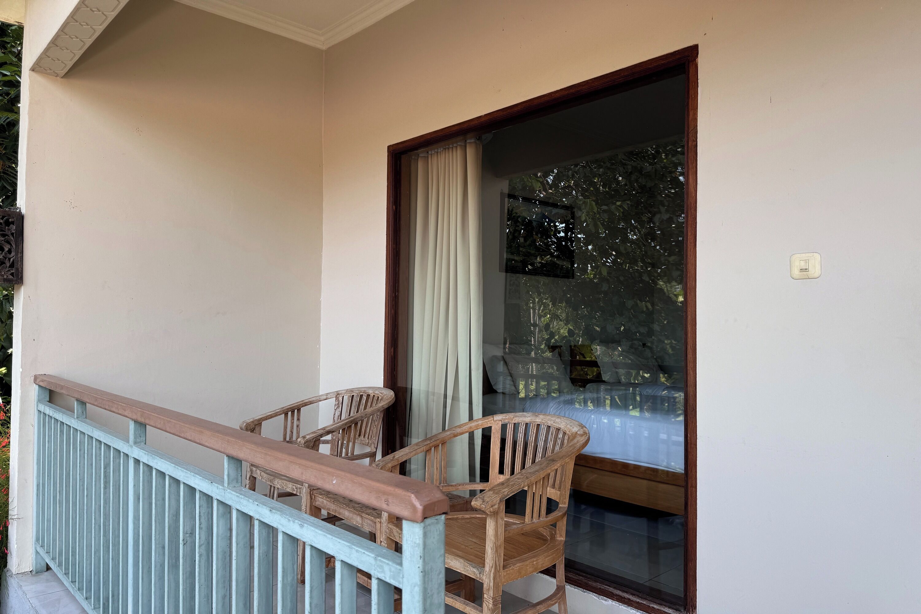 Ein Balkon mit einem Holzstuhl und einem Tisch, eine Glastür mit Vorhang und Blick auf grüne Vegetation.