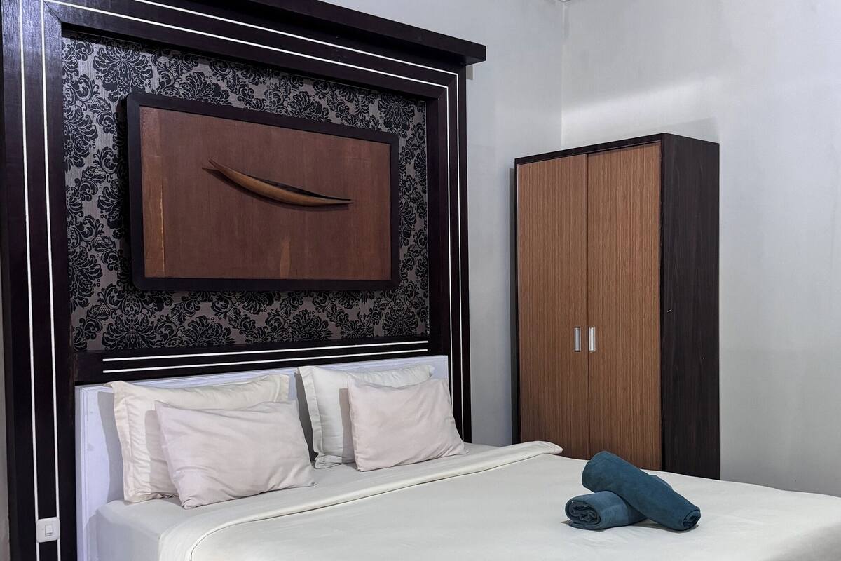 A neatly made bed with a white mattress and pillows, a wooden headboard with a decorative panel, and a wardrobe with two doors.