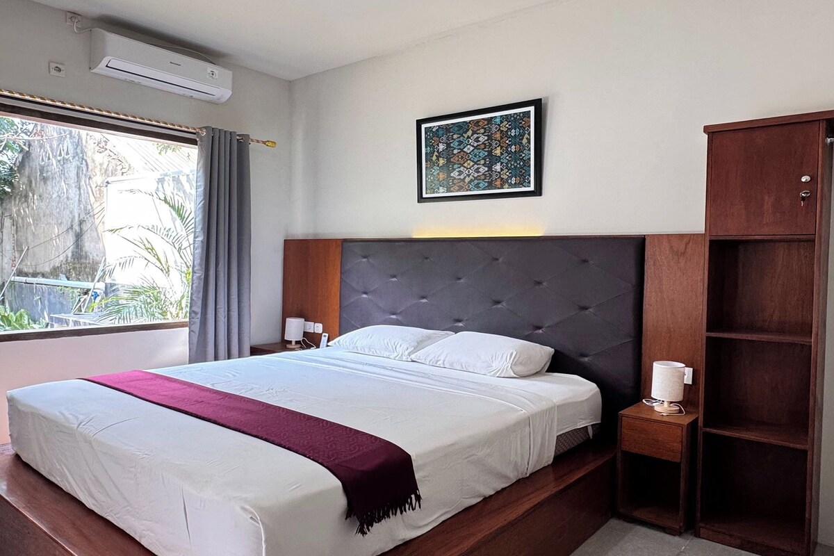 Un dormitorio con una cama grande, una mesita de noche, un armario de madera, una lámpara en el techo y una ventana con cortinas.