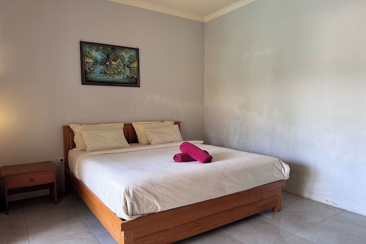 A neatly made bed with a pink towel, a wooden nightstand, and a painting on the wall.