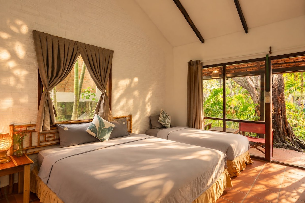 A room with two beds, a window with curtains, a view of greenery outside, and a wooden ceiling beam.