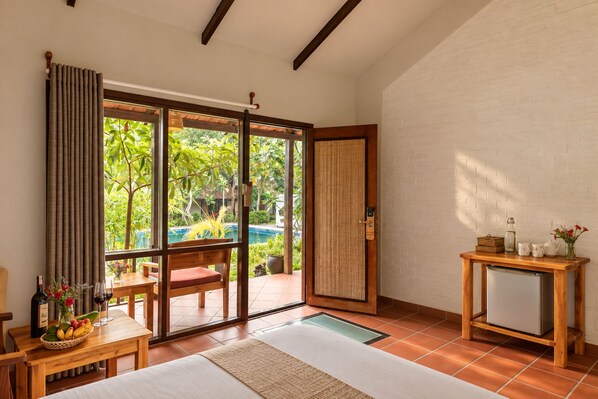 A room with a sliding glass door leading to a pool, a wooden table with a bottle and fruit, and a small table with a vase and a bottle. - An Nhien Retreat Phu Quoc (Phu Quoc)
