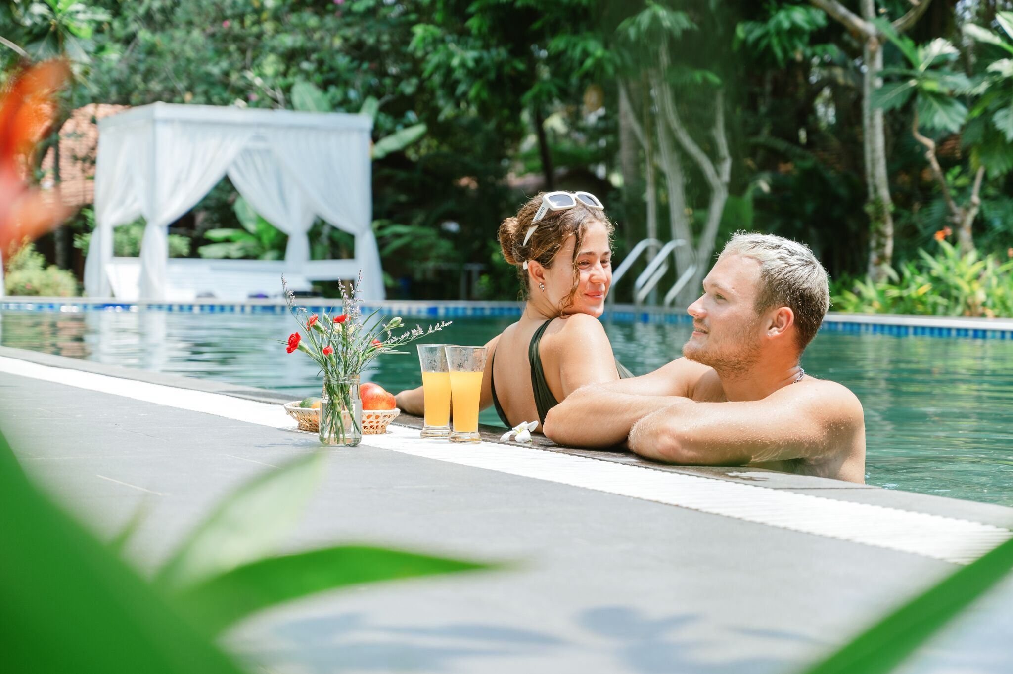 Una pareja descansando junto a una piscina, con una mesita que sostiene bebidas y un florero con flores.
