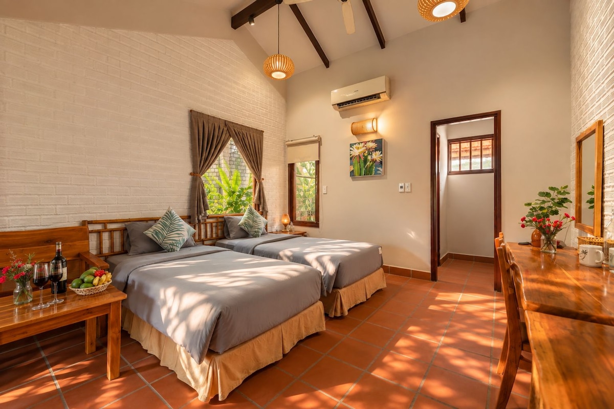 A bedroom with two beds, a wooden bench, a dining table, and a large window with curtains.