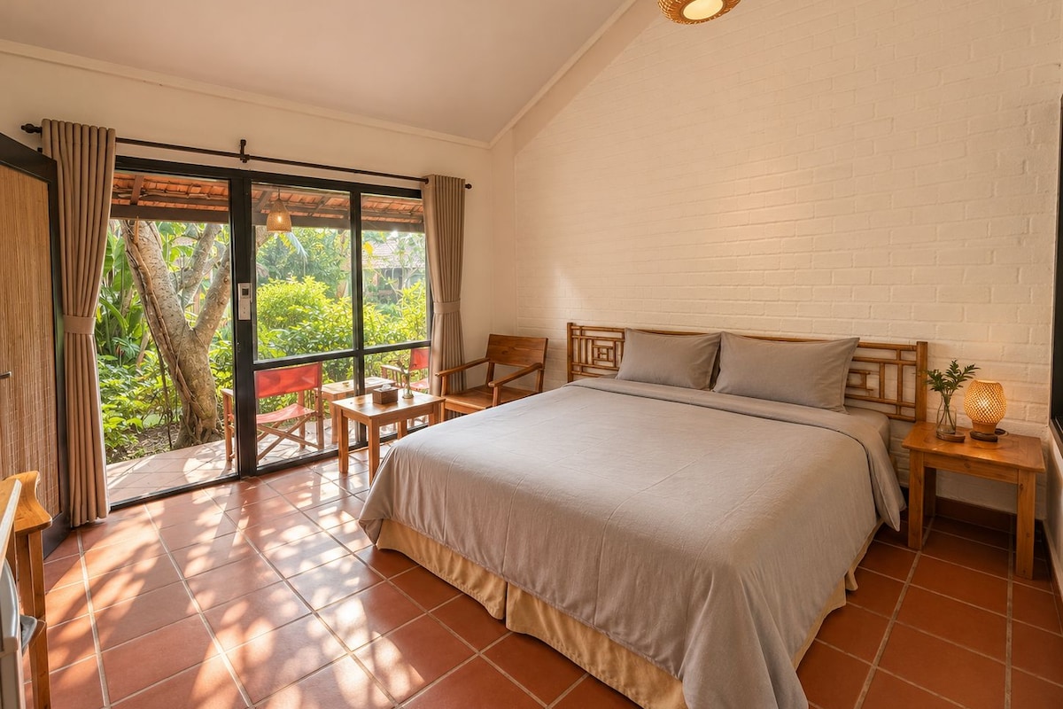 A bedroom with a large bed, a wooden nightstand, a small table, and a chair. There is a sliding glass door leading to an outdoor area with a view of greenery.