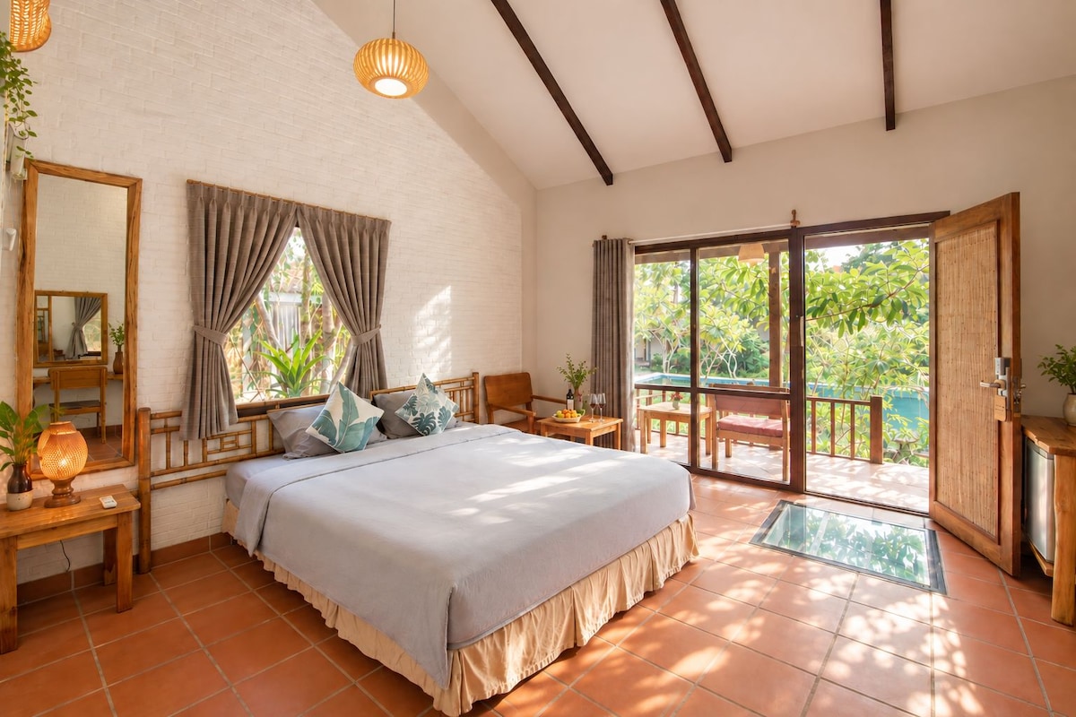 A bedroom with a bed, bedside tables, a mirror, and a view of a pool and greenery.