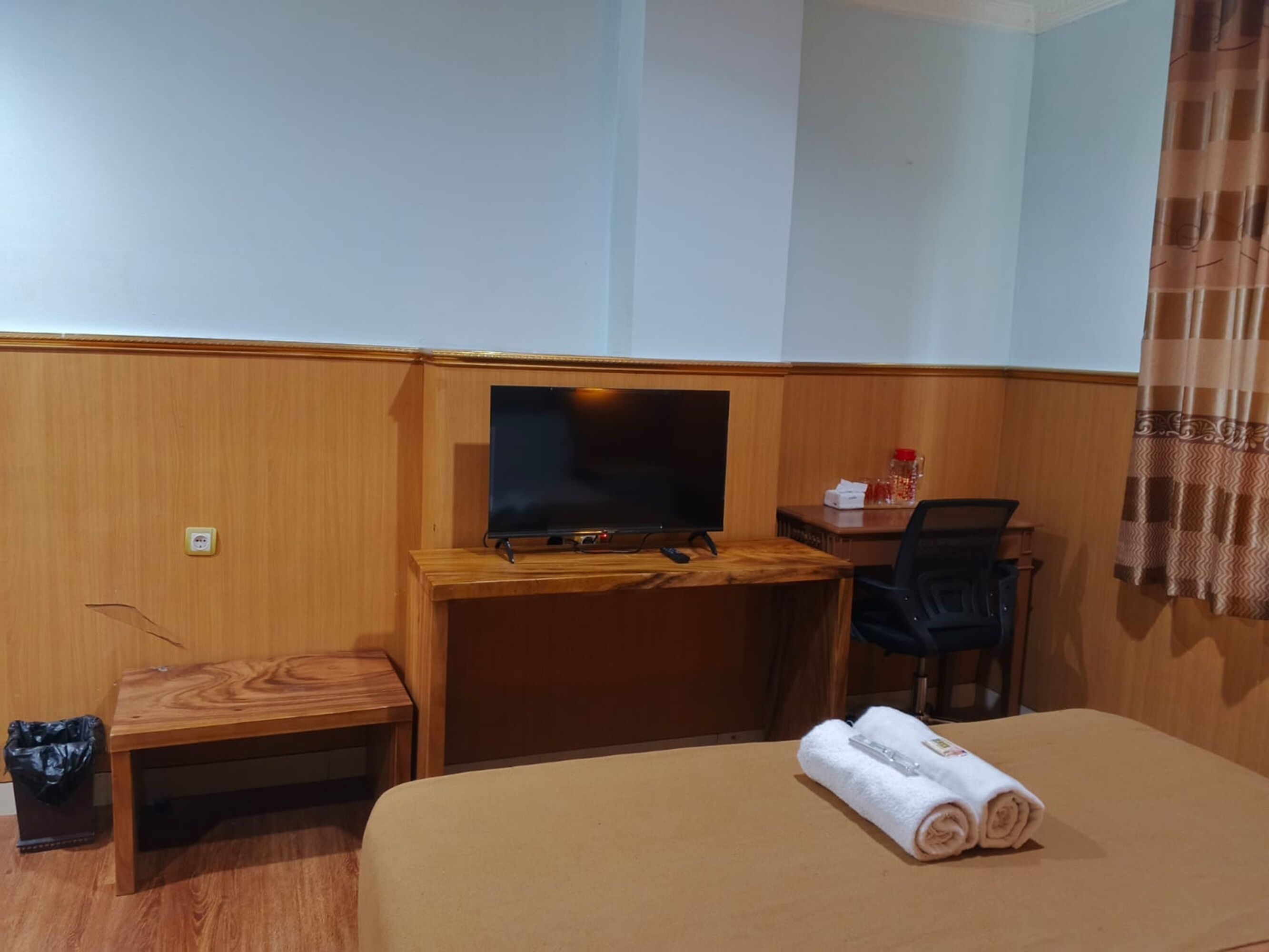 A hotel room with a wooden desk, a television, a chair, and a bed with rolled towels.