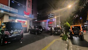 Escena callejera con varios vehículos y tiendas, incluyendo un hotel y un bar.