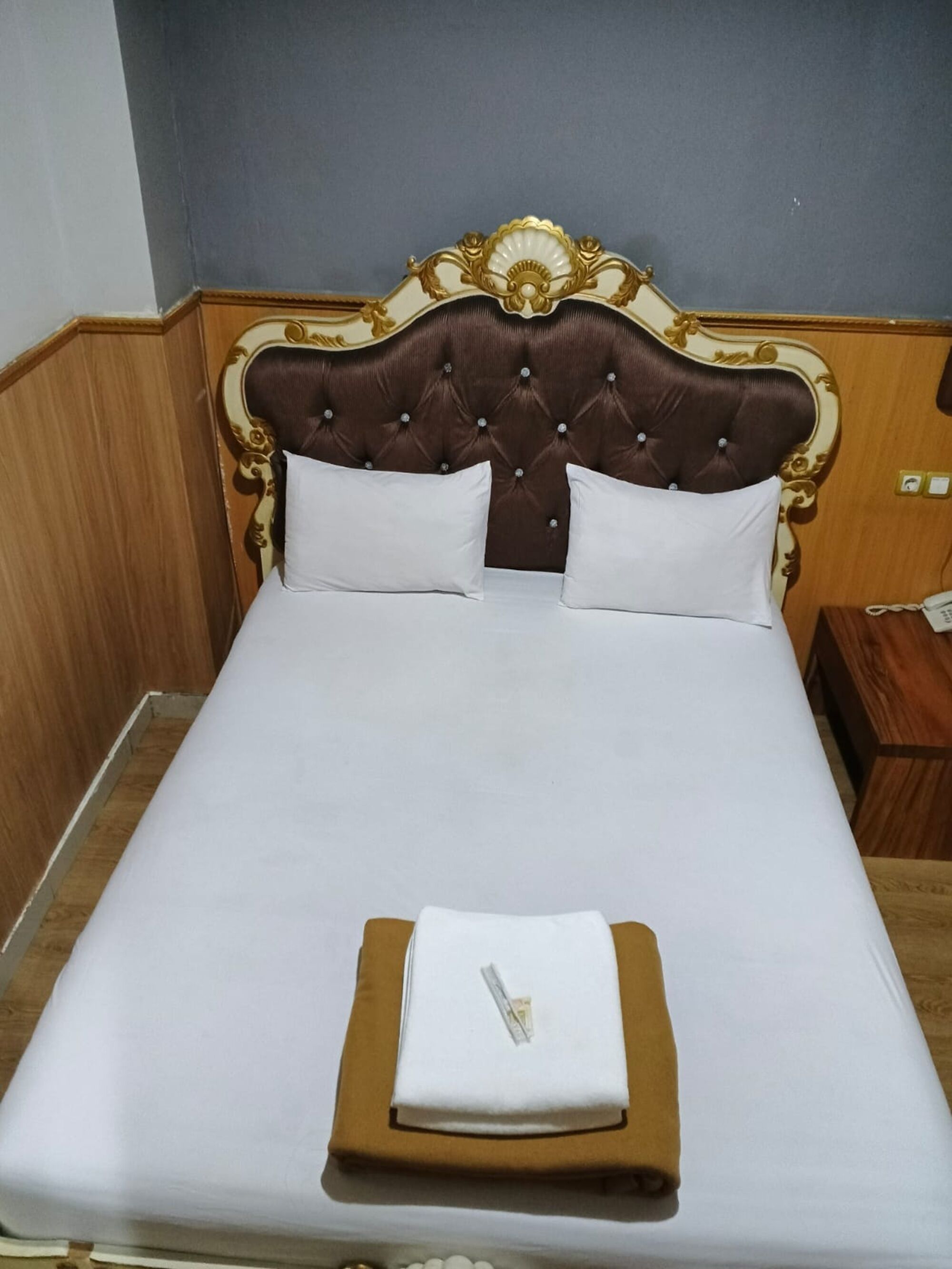 Una cama bien tendida con un cabecero capitonado, ropa de cama blanca y una toalla doblada sobre una mesita de noche de madera.