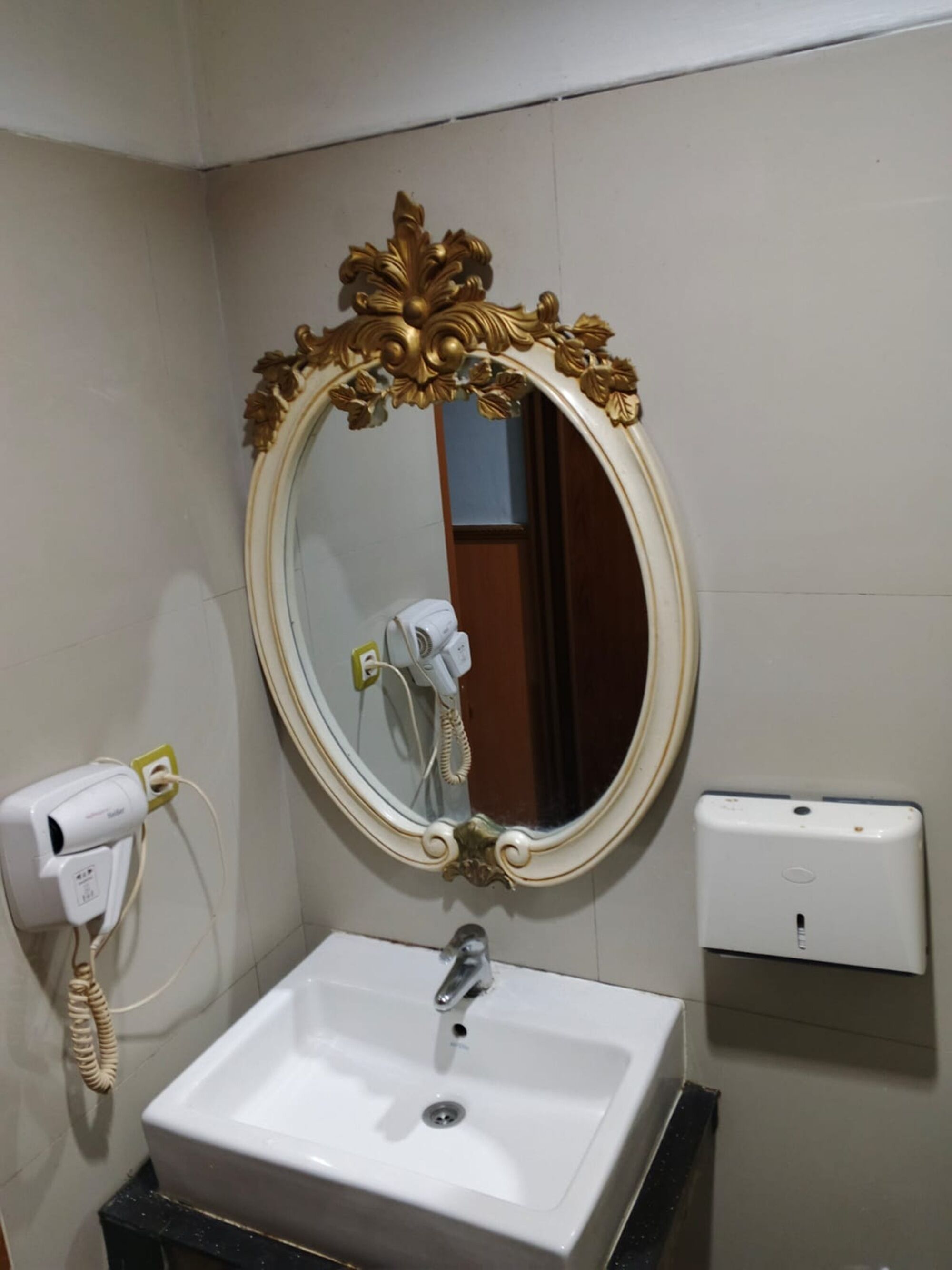 A bathroom with a white sink, a gold-framed mirror, a hairdryer, and a soap dispenser.