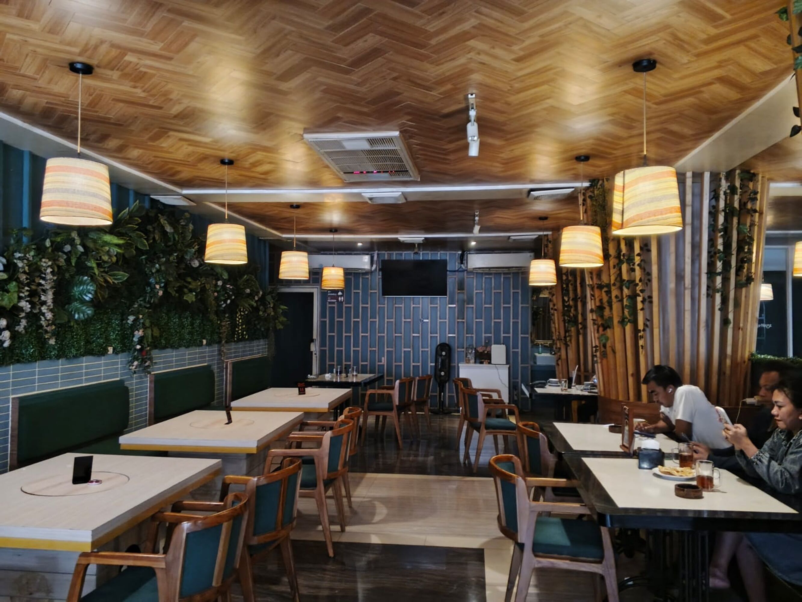 A restaurant with wooden ceiling and walls, green plants, and striped pendant lights. Tables and chairs are arranged for dining.