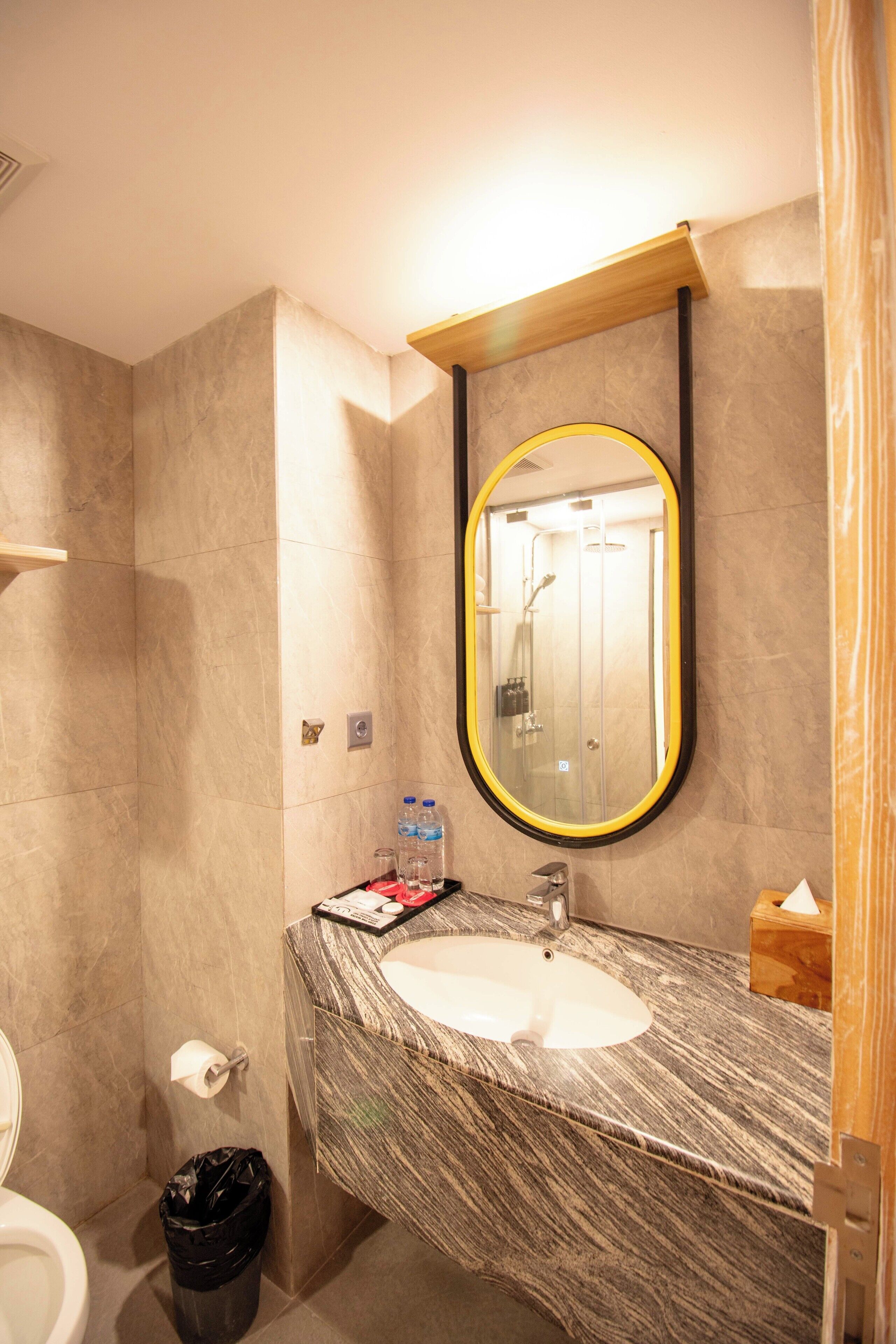 Un baño con lavamanos de mármol, un espejo con marco amarillo y una ducha que se ve en el reflejo.