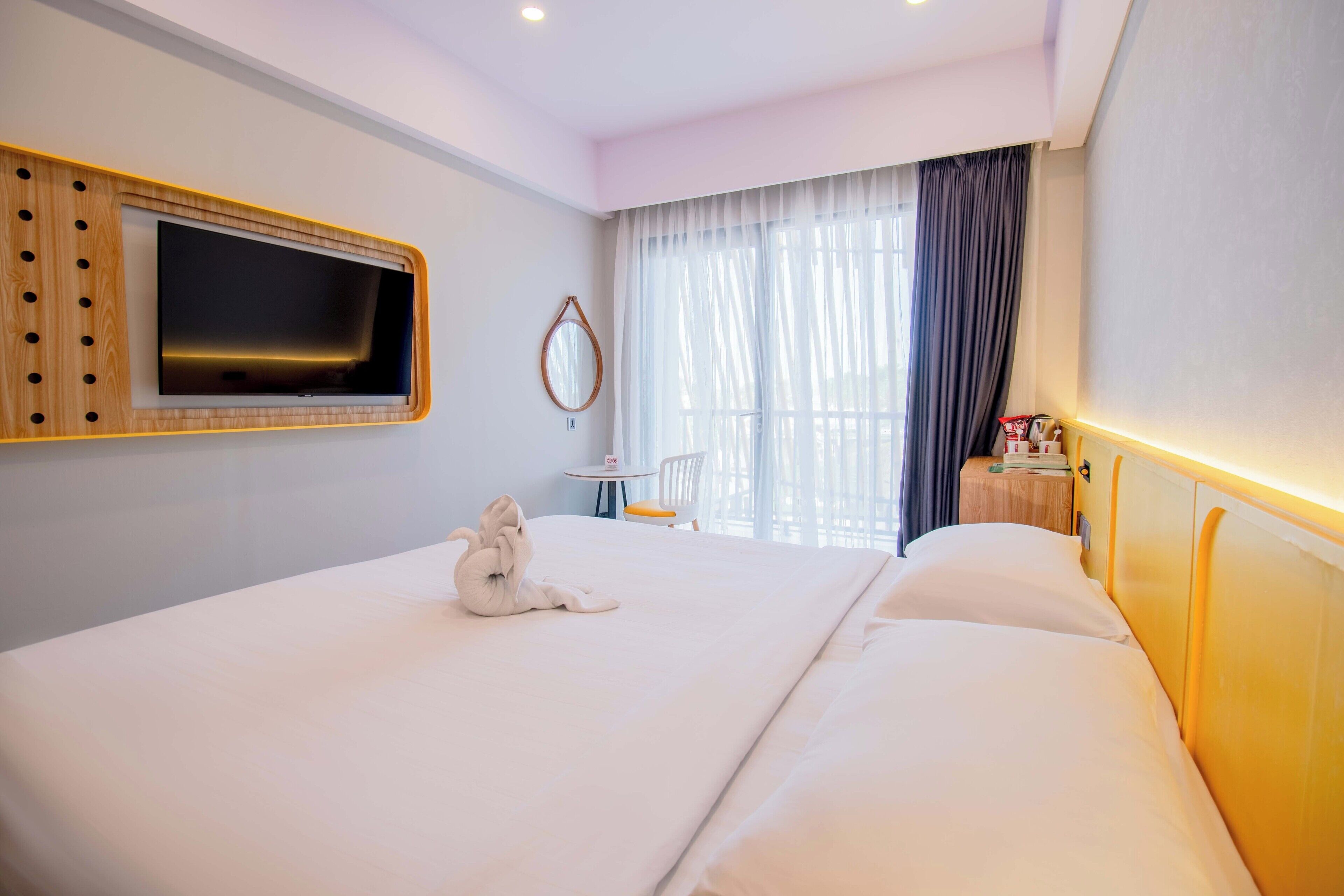 Habitación de hotel con una cama grande, un televisor de pantalla plana montado en la pared, una mesita redonda con una silla y una ventana con cortinas.