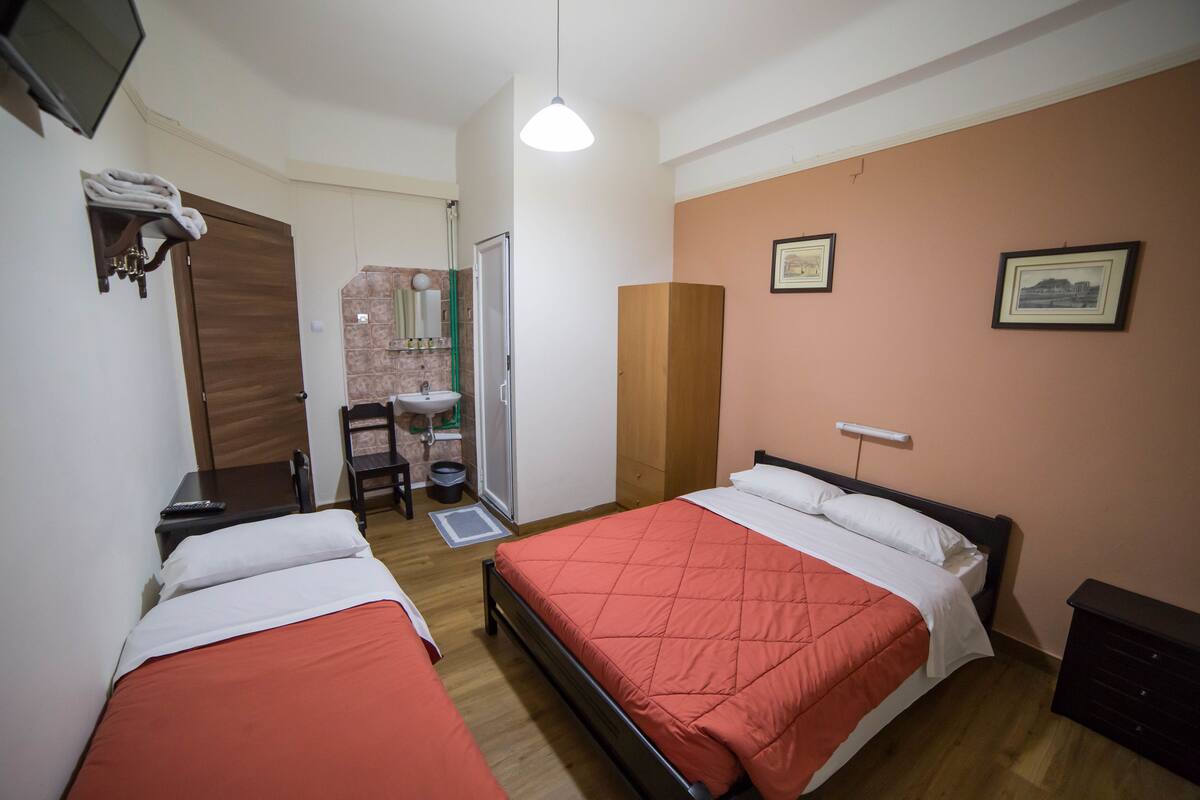A bedroom with two beds, a wooden wardrobe, a desk with a chair, a sink, and a mirror.