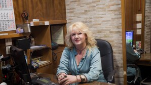 A woman sitting at a desk in an office with a brick wall, a calendar, and various office equipment. - Tempi Hotel (Athens)