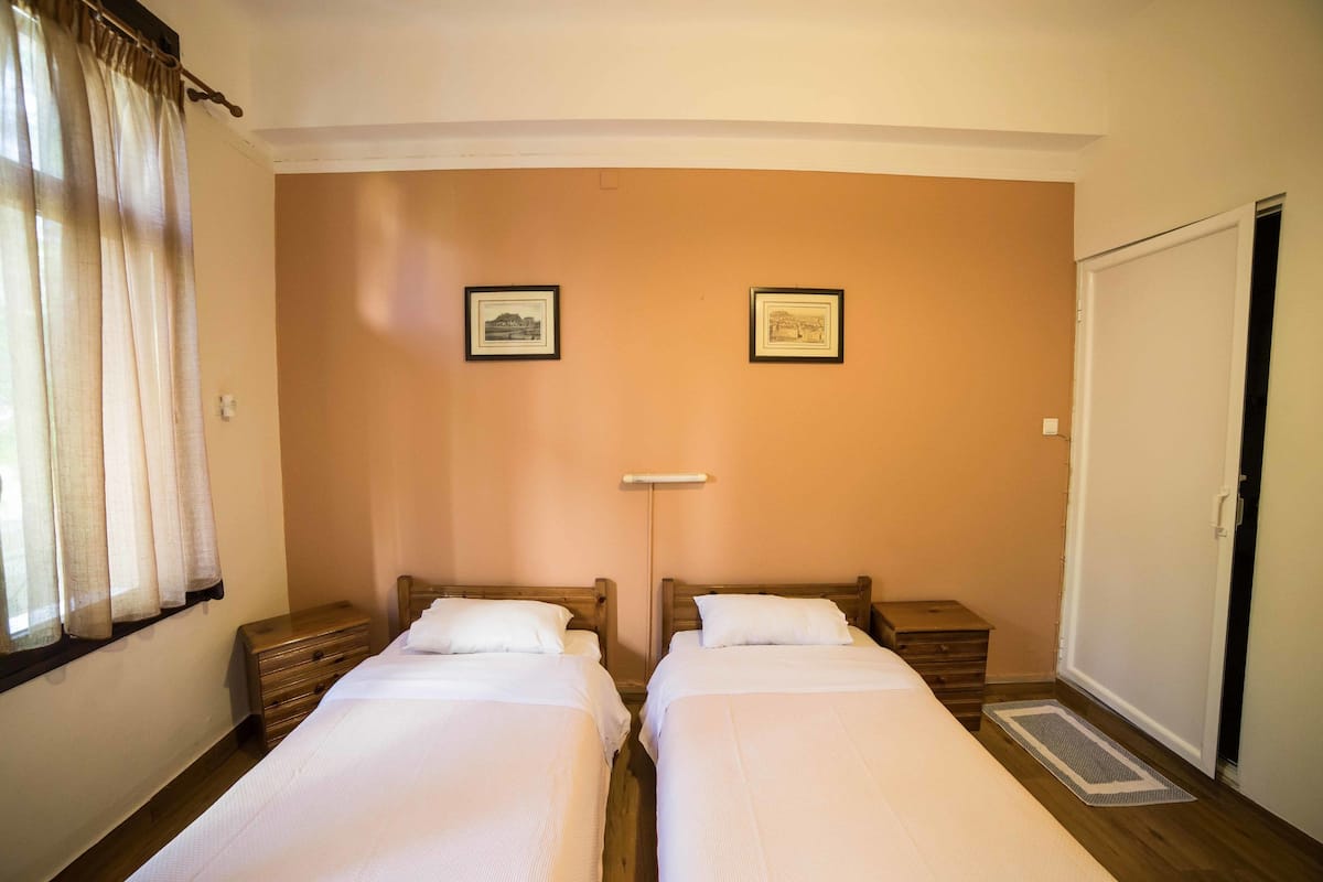 A bedroom with two single beds, a bedside table with a lamp, a wardrobe, and a window with curtains.