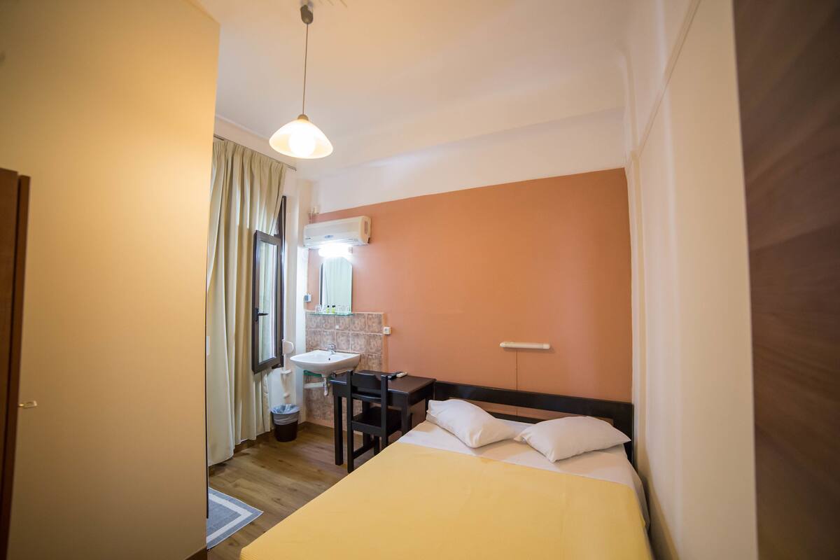 A bedroom with a bed, a desk, a chair, a window, a sink, and an air conditioning unit.