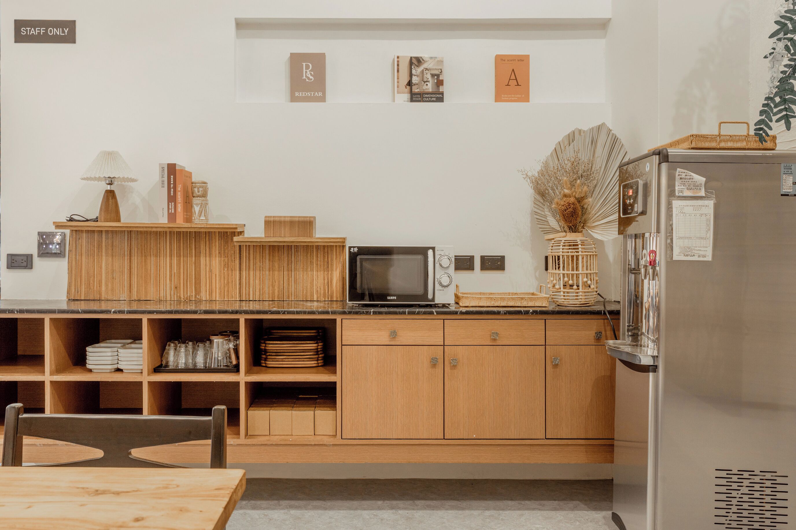 A modern kitchen with wooden cabinets, a microwave, and a refrigerator. There are shelves with dishes and a table with chairs.