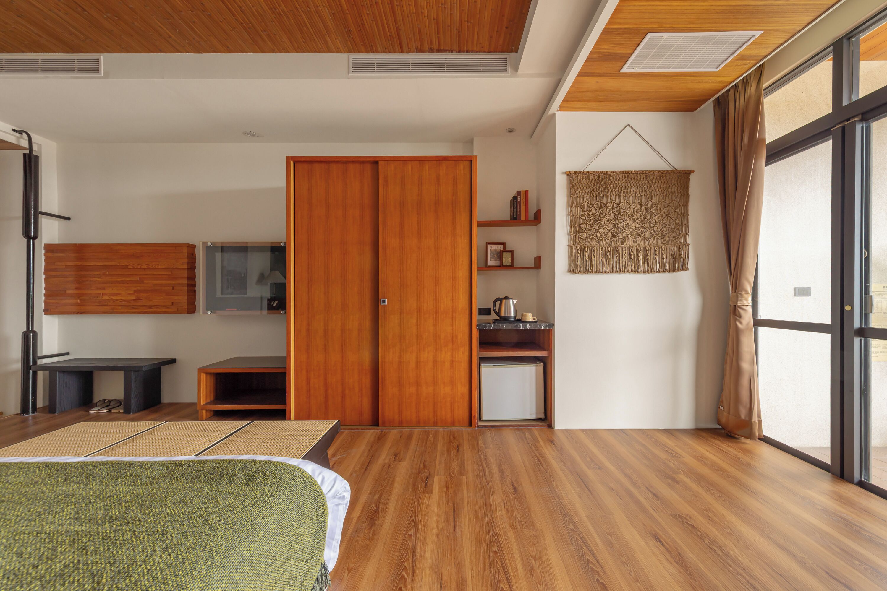 A modern room with a wooden floor, a bed, a TV, a wardrobe, a kitchenette, and a large window with curtains.