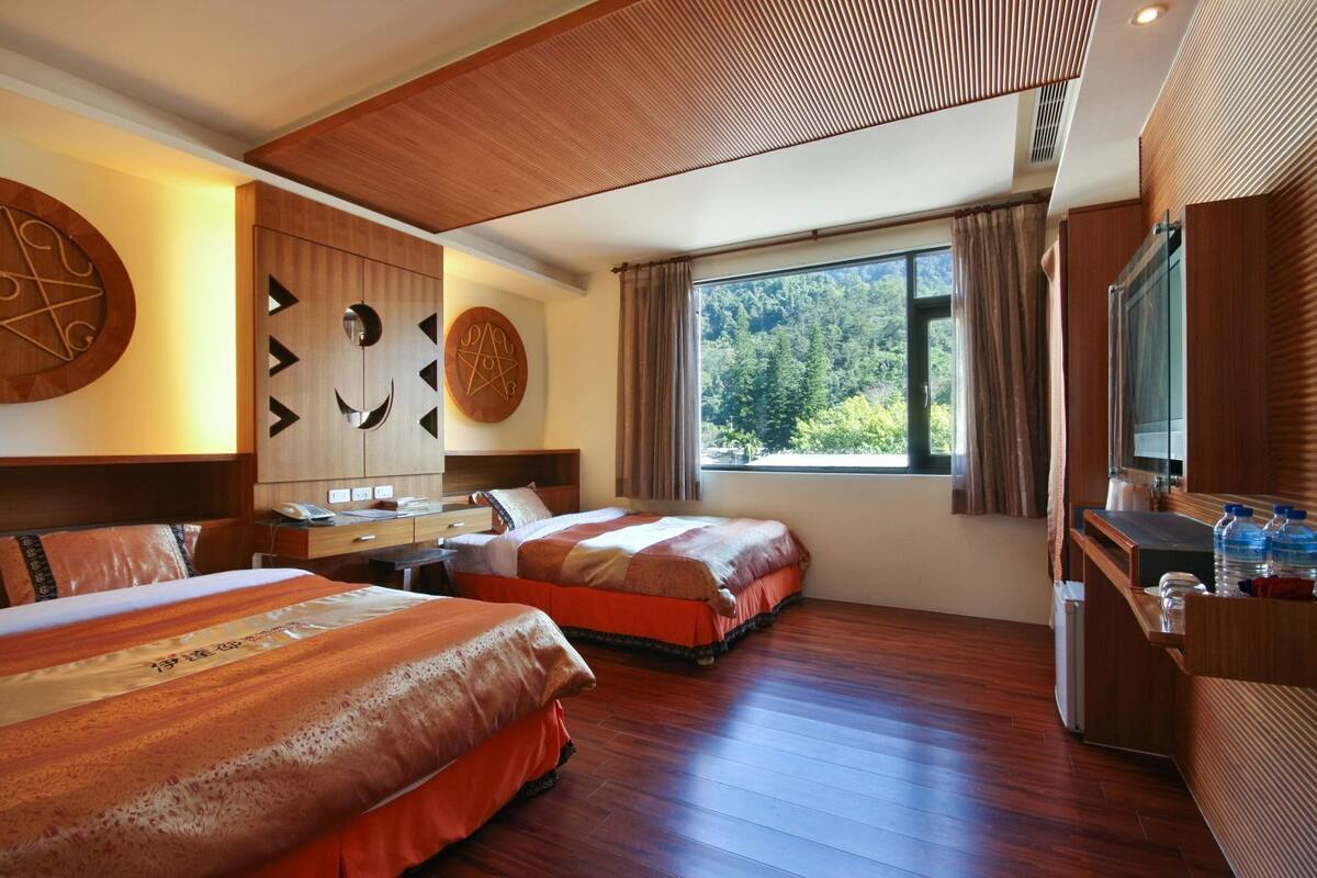 Une chambre d’hôtel avec deux lits, un bureau, un miroir, une fenêtre donnant sur des arbres et un plafond en bois.
