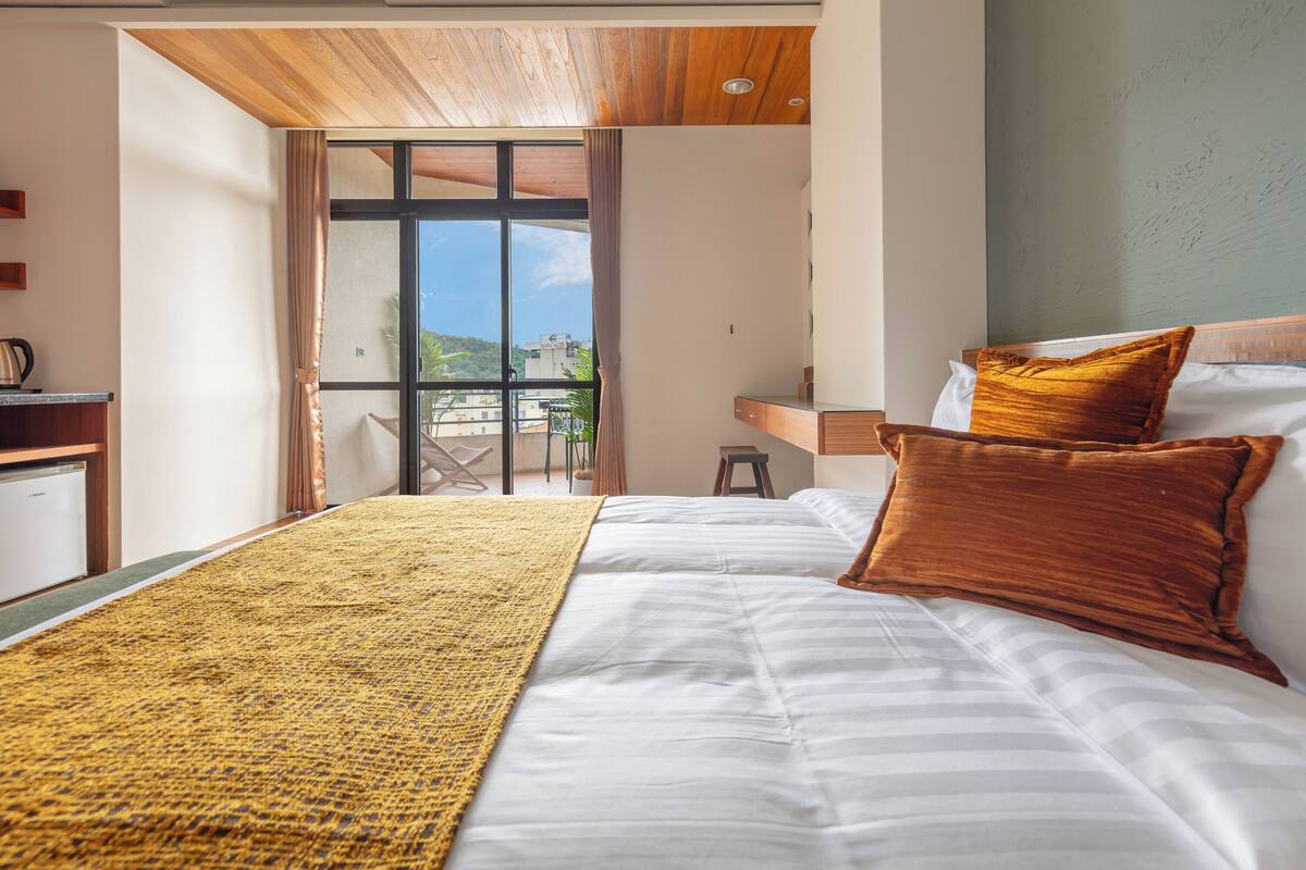 Une chambre d’hôtel avec un lit, un plafond en bois, une vue à travers une porte vitrée et un bureau avec une lampe.
